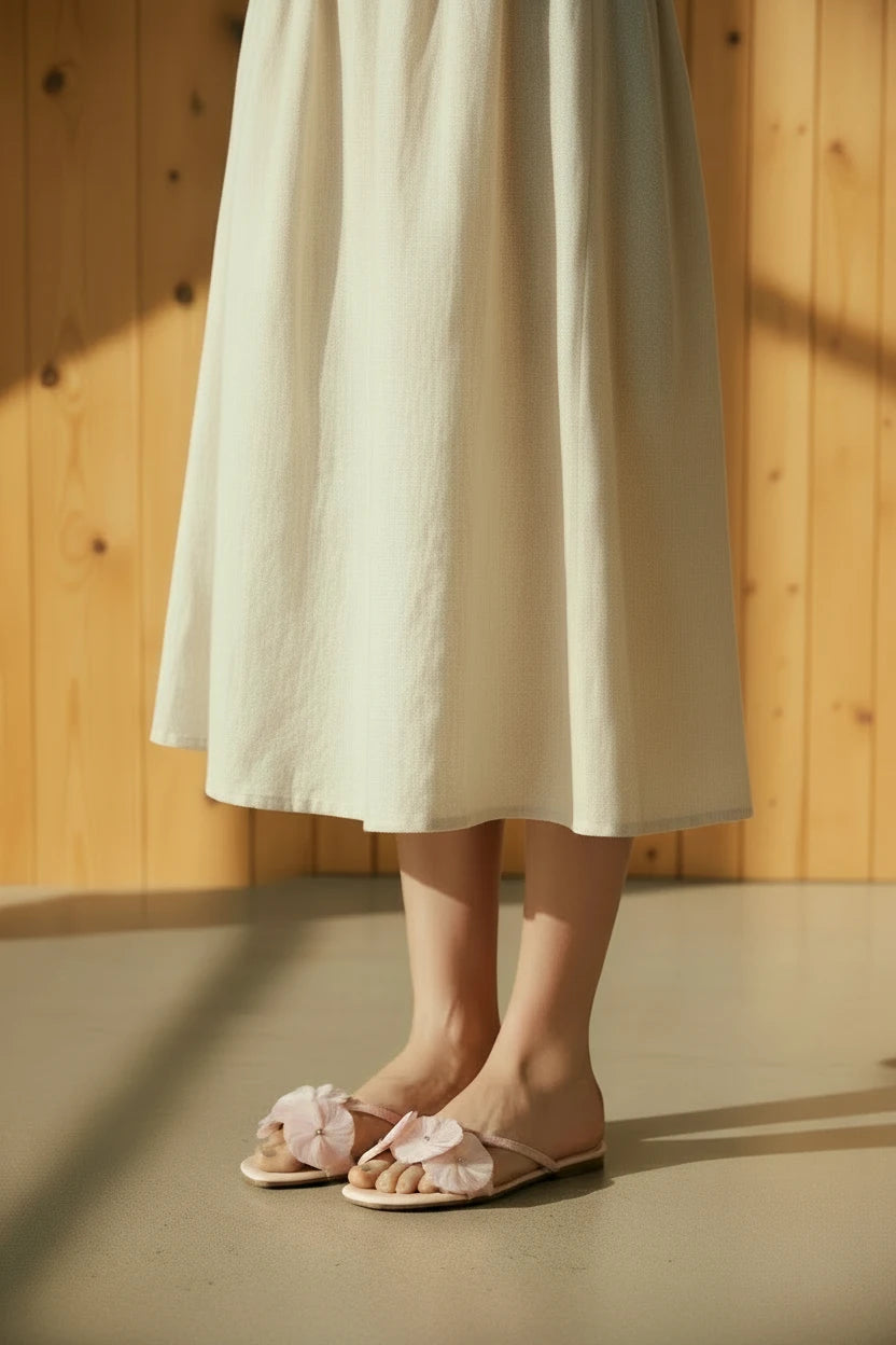 Primer plano de sandalias planas MD en color rosa pastel con detalles de flores textiles sobre suelo de microcemento y fondo de madera.